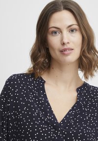 Marinebluse mit weißen Punkten, V-Ausschnitt und kurzen Ärmeln. Leichtes Gewebe mit glatter Textur. Lässiges Design, das für verschiedene Anlässe geeignet ist.