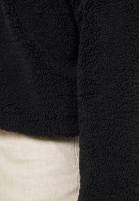 Czarna kurtka z polaru o miękkim, teksturowanym wykończeniu, z krótkim krojem i widocznym przeszyciem. Nosi się ją na jasnym materiale.