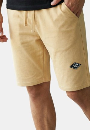 Homme portant un short beige jusqu'aux genoux avec une taille à cordon et un petit logo noir en forme de losange sur la jambe gauche.
