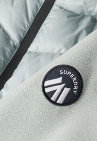 Tejido de forro polar verde claro con una textura suave, que presenta un parche redondo negro con el logo blanco "Superdry" y acentos de diseño geométrico.