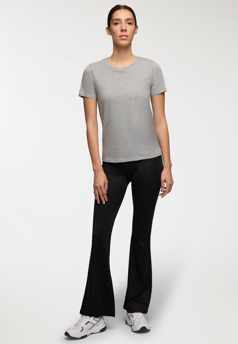 Szary t-shirt z krótkim rękawem w połączeniu z czarnymi spodniami w dzwony. Spodnie mają gładką teksturę, a model ma na sobie białe buty sportowe.