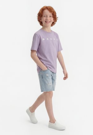 Χαμογελαστός κοκκινομάλλης αγόρι που περπατά με τα χέρια στις τσέπες, φοράει λιλά μπλούζα, ανοιχτόχρωμα τζιν σορτς με φθορές και λευκά παπούτσια slip-on.