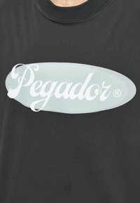 Černé bavlněné tričko s velkým světlým šedým oválem, na kterém je text "Pegar" v bílém kurzívním písmu. Hladký materiál s kulatým výstřihem.