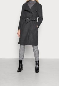 Harmaa villasekoitustakki käärittävällä mallilla, kauluksella ja mustilla keinonahkakoristeilla. Päällä ruutukuvioiset housut ja mustat nilkkurit.