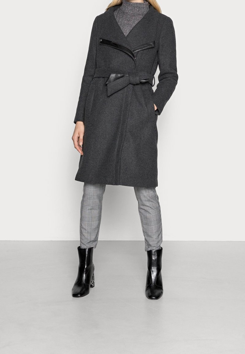 Harmaa villasekoitustakki käärittävällä mallilla, kauluksella ja mustilla keinonahkakoristeilla. Päällä ruutukuvioiset housut ja mustat nilkkurit.