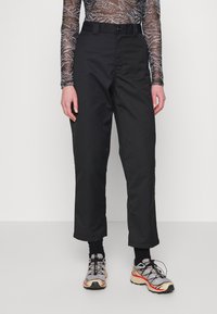 Black trousers featuring a straight cut, belt loops, and a button closure. Worn with gray and black patterned mesh top and sporty shoes.