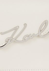 Sac à main en cuir blanc avec un logo en cursive argenté "Karl" bien en évidence sur le devant, accentué par une texture lisse et des détails cousus.