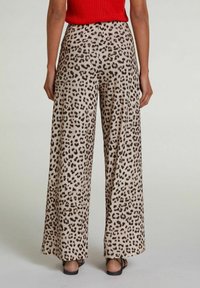Person iført bredbenede bukser med brun og sort leopardprint, rød top og sorte sandaler, stående foran en ensfarvet baggrund.