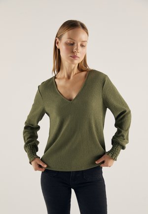 Olive green textured sweater with a V-neck and long sleeves featuring gathered cuffs. Worn over dark jeans, it has a loose fit.