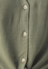 Grüne Hemdbluse aus strukturiertem Stoff mit fünf cremefarbenen Knöpfen entlang der Vorderseite. Schlichtes und sauberes Design.
