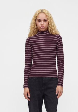 Joven mujer con cabello rubio rizado que lleva un jersey de cuello alto a rayas granate y blancas y pantalones negros, de pie frente a un fondo liso.