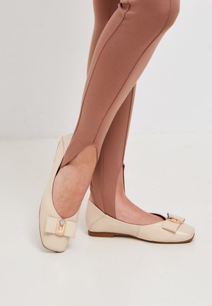 Ballerines beiges avec un accent en nœud et un matériel en gemme. Matériau en cuir lisse, bout rond, talon bas, accompagnées de leggings marron ajustés.
