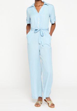 Person iført lyseblå jumpsuit med knapper, bundet taljebælte og opkørte ærmer, kombineret med flettede slip-on sandaler.