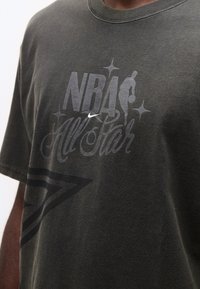 Κοντινό πλάνο σε σκούρο γκρι μπλουζάκι με το λογότυπο NBA All Star, το σήμα Nike και γραφικό με μπασκετμπολίστα που κρατάει μπάλα.