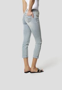 Világoskék, testhezálló, bokáig érő farmer nadrág felhajtott szegélyekkel, hátsó zsebek varrási részletekkel, fekete papucs szandálokkal párosítva.