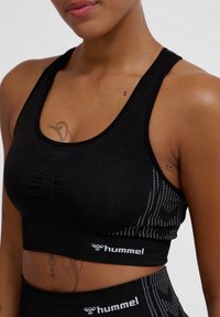 Svart sport-bh med ett strukturerat tyg, med en samlad mitt och en vit logotypdetalj i fållen. Sidopanelen visar ett prickigt mönster.