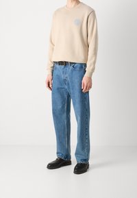 Persona che indossa un felpa beige, jeans blu con una cintura nera e mocassini neri, in piedi di fronte a uno sfondo bianco semplice.