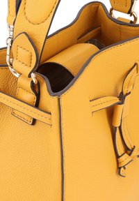 Sac seau en cuir jaune avec finition texturée, garniture sombre contrastante et liens décoratifs sur les côtés. Bretelle réglable incluse.