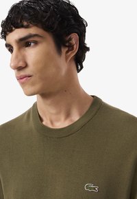 Joven con cabello rizado y oscuro, vistiendo un suéter olivo de Lacoste, mirando ligeramente hacia la izquierda sobre un fondo blanco liso.
