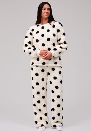 Jeune femme debout portant un ensemble de pyjama blanc duveteux avec de gros pois noirs et des chaussons assortis sur un fond uni.