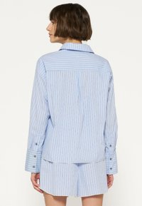 Camisa a rayas azul claro con mangas largas y puños con botones, que presenta un canesú en la parte posterior y un sutil detalle de logo en la parte superior de la espalda.