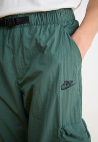 Green, lightweight pants with a wrinkled texture, elastic waistband, black buckle, and a black Nike logo on the left thigh.