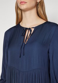 Ναυτικό μπλε πουκάμισο με δεξιά κολάρο, μακριά μανίκια και πτυχωτό σχέδιο. Λείες υφή με απαλό γυαλίσμα, δημιουργώντας μια ρομαντική σιλουέτα.