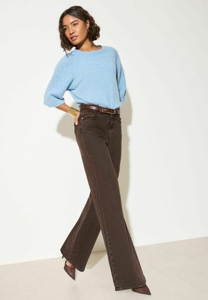 Woman wearing a light blue sweater, brown wide-leg jeans, brown heels and a gold cuff bracelet, posing against a light background.