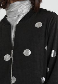 Giubbotto bomber nero con grandi pois metallici argento, chiusura a zip sul davanti e colletto a coste. Tessuto morbido e texture.