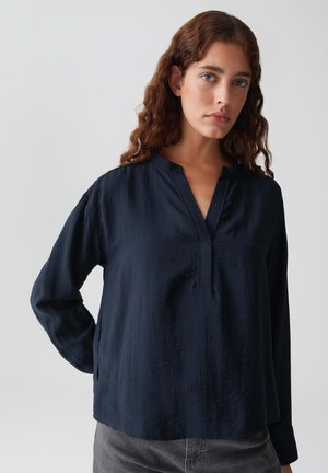 Vrouw met golvend bruin haar, gekleed in een lang mouw, donkerblauwe blouse en grijze broek, staande tegen een effen lichte achtergrond.