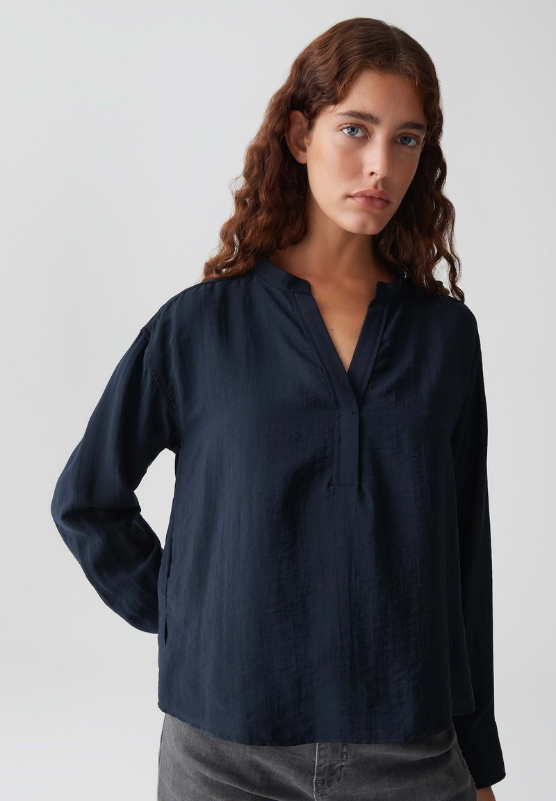 Una donna con capelli castani mossi che indossa una blusa a maniche lunghe di un blu navy scuro e pantaloni grigi, in piedi contro uno sfondo chiaro e uniforme.