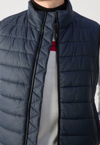 Granatowa kamizelka puchowa bez rękawów z wysokim kołnierzem, pikowaną teksturą i zapięciem na zamek, założona na jasną koszulę w poziome paski.