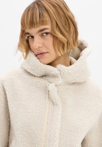 Crèmekleurige hooded jack van gestructureerde fleece, met een hoge kraag en een sluiting met een middensteentje. Gladde voering zichtbaar bij de randen.