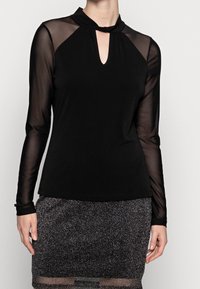 Blusa nera a maniche lunghe con scollatura a chiave, maniche trasparenti a contrasto e una texture di tessuto morbido, abbinata a una gonna nera scintillante.