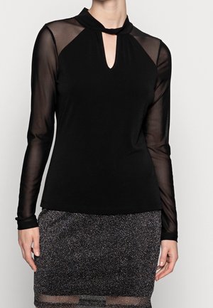 Blouse noire à manches longues avec un décolleté en trou de serrure, des manches transparentes contrastantes et une texture de tissu douce, associée à une jupe noire scintillante.