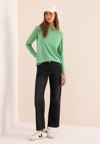 Hellgrünes Langarmoberteil kombiniert mit schwarzen weiten Jeans und weißen Sneakern mit schwarzen Akzenten. Eine schlichte Mütze rundet das Outfit ab.