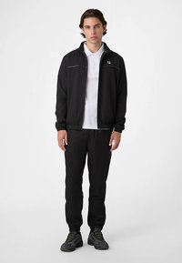 Sort zip-up jakke med elastiske manchetter, hvid polo under, sorte bukser og mørke sneakers. Minimalistisk design med logodetalje.