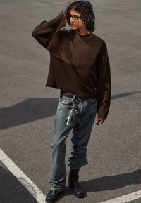 Personne portant un pull oversize marron, un jean bleu avec un porte-clé, des bottes noires, des lunettes et des écouteurs, se tenant sur de l'asphalte.