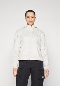 Giacca con cappuccio di colore crema chiaro, con zip frontale, polsini elastici e due tasche laterali. Presenta il logo sul lato sinistro superiore.