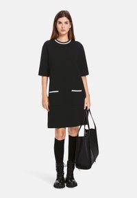 Schwarzes Kleid mit kurzen Ärmeln und Rundhalsausschnitt, zwei Fronttaschen, weißen Besätzen, kombiniert mit kniehohen Socken und schwarzen Schnürstiefeln, während sie eine Tasche hält.
