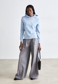 Ανοιχτό μπλε φούτερ με λευκό λογότυπο, σε συνδυασμό με γκρι παντελόνι φαρδιάς εφαρμογής με ρίγες. Μαύρη τσάντα ώμου προσθέτει αντίθεση. Απαλές υφές και χαλαρή εφαρμογή.