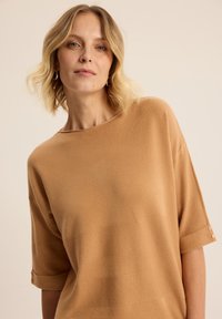 Beżowy sweter o luźnym kroju, z krótkim rękawem i poziomą teksturą rib, ozdobiony guzikami na mankietach.