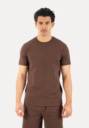 Mężczyzna stojący przodem, ubrany w gładki brązowy t-shirt z krótkim rękawem i pasujące brązowe spodnie na neutralnym tle.