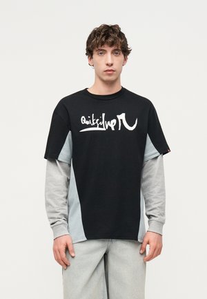 Νέος άνδρας με μαύρο και γκρι μακρυμάνικο T-shirt σε στρώσεις με λευκό γραφικό κείμενο, στέκεται μπροστά σε ένα απλό φωτεινό φόντο.