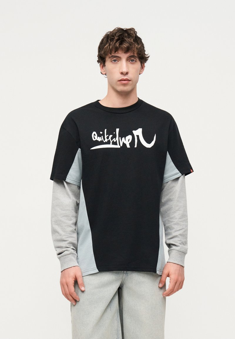 Νέος άνδρας με μαύρο και γκρι μακρυμάνικο T-shirt σε στρώσεις με λευκό γραφικό κείμενο, στέκεται μπροστά σε ένα απλό φωτεινό φόντο.