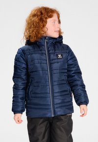 Marineblauwe pufferjack met horizontale stiksels, ritssluiting en capuchon. Heeft een klein logo en twee zijzakken. Draagt over donkere broeken.