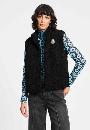 Vrouw die een zwart gestructureerd vest draagt met een bloembroche over een blauw-zwart gebloemde top met lange mouwen en donkergrijze broek, staand tegen een witte achtergrond.
