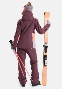Ski outfit in dark burgundy with pink and gray accents. Features waterproof jacket, pants, gloves, and carries skis and poles. Textured beanie.