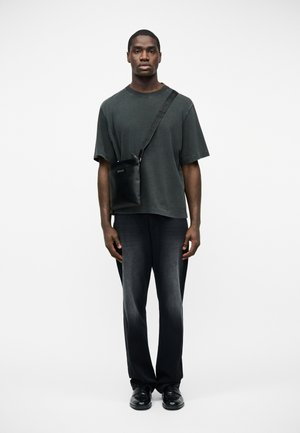 Młody mężczyzna w wyblakłej czarnej, luźnej koszulce, ciemnych wyblakłych dżinsach, czarnych butach i czarnej skórzanej torbie przewieszonej przez ramię, stojący na białym tle.