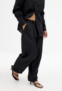 Črna lanena srajca in usklajen hlač s prostornim krojem, z stransko žepom in diskretno obrobo. V kombinaciji s črnimi sandalarmi na peto.